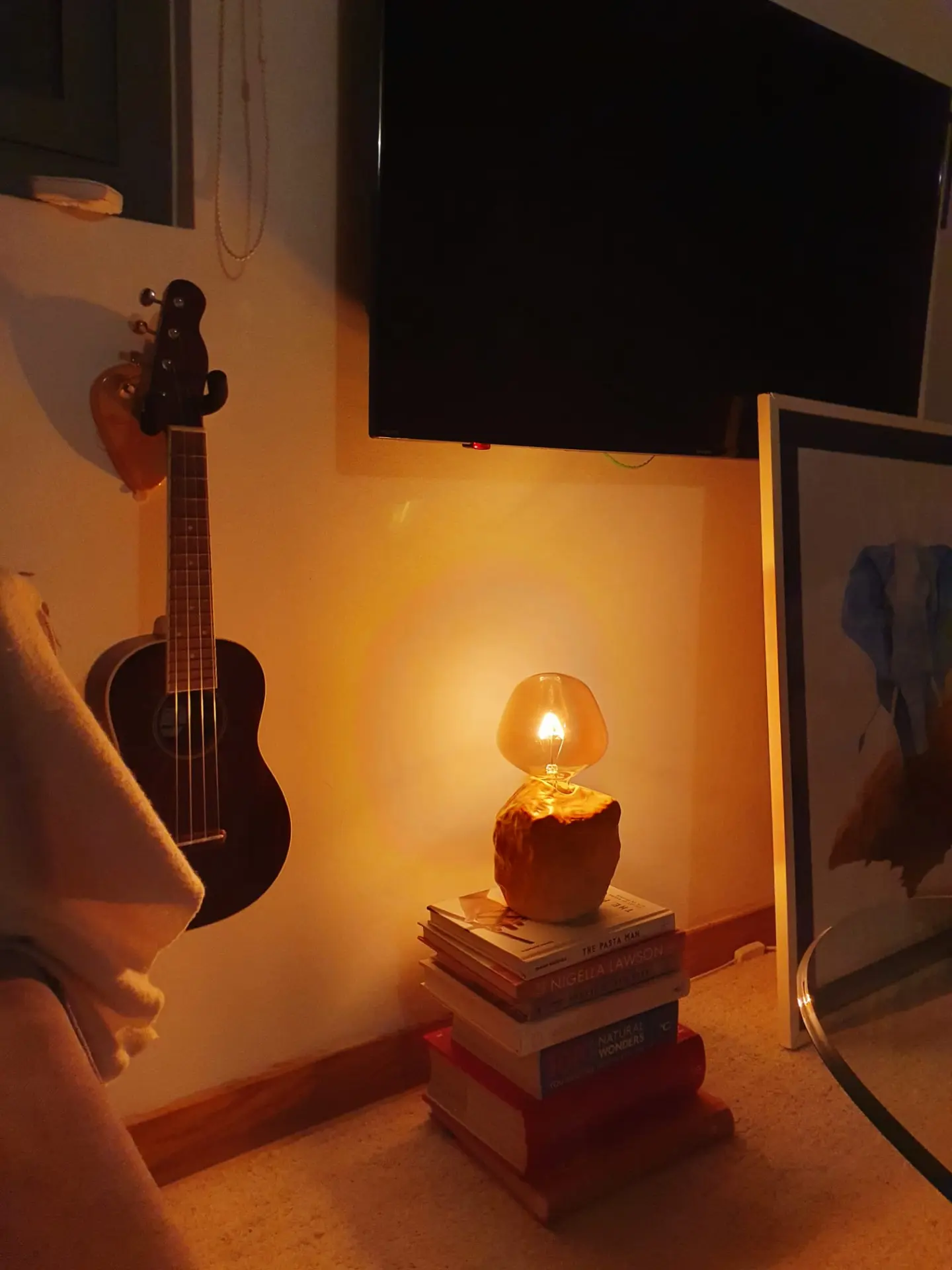 Small ceramic lamp glowing warmly on a stack of books beside a ukulele