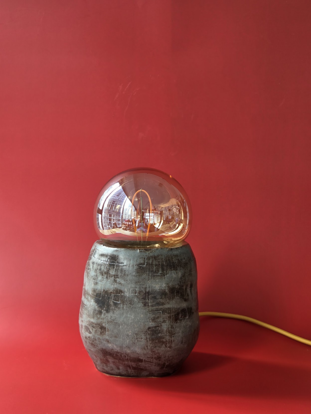 Tarn, an irregular dark stoneware table lamp topped with a warm glass globe against a red backdrop