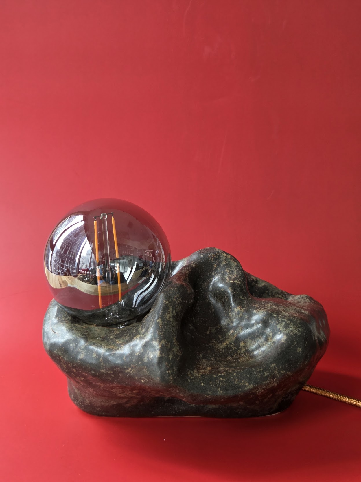 Shoal, a dark mottled stoneware table lamp with a folding, wave-like form and smoked glass globe against a red backdrop