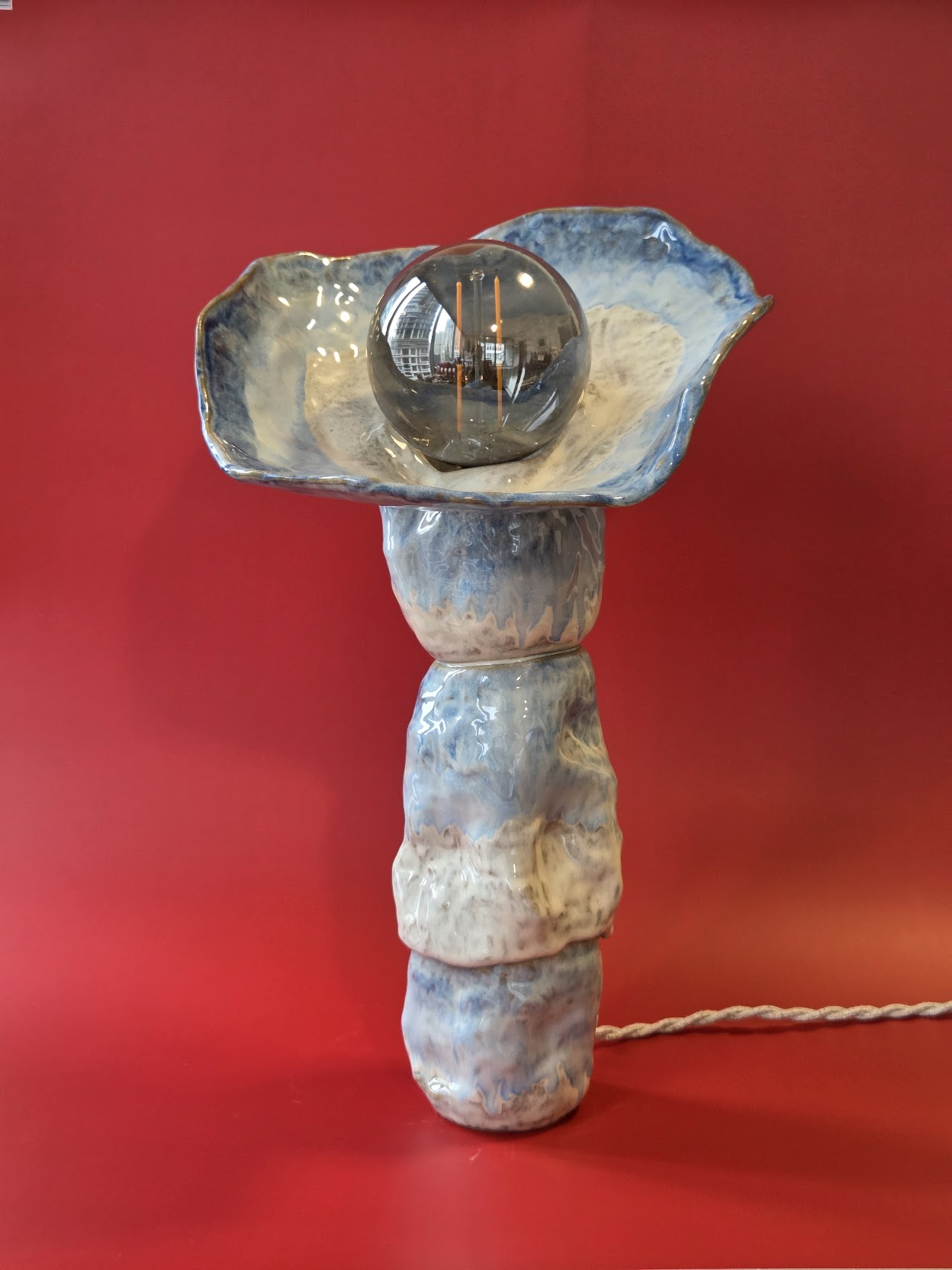 Riffle, a stacked blue and cream glazed stoneware table lamp with a flared top form and smoked glass globe against a red backdrop