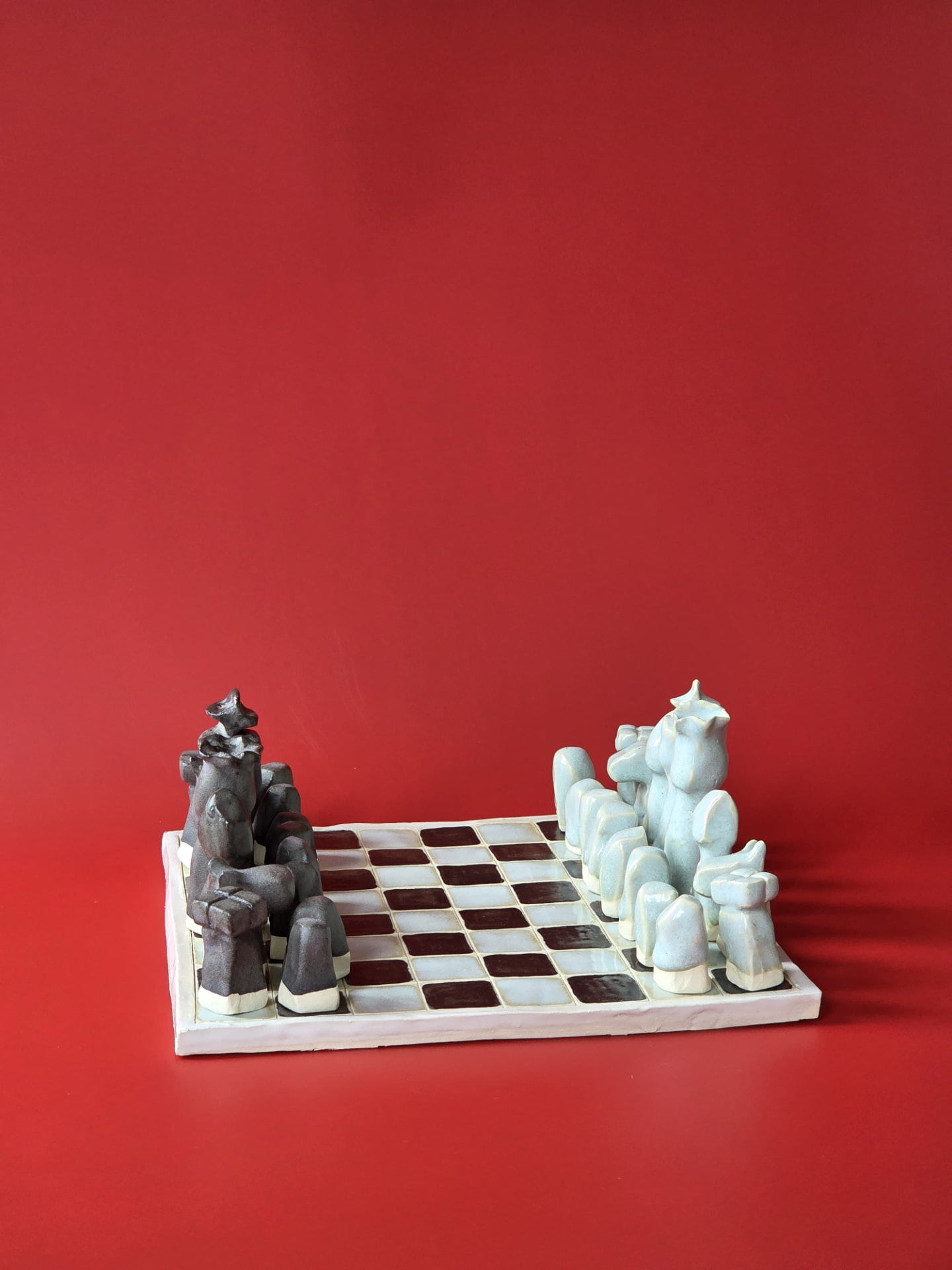 Littoral, a handbuilt ceramic chess set with eroded, stone-like pieces on a glazed checkered board against a red backdrop