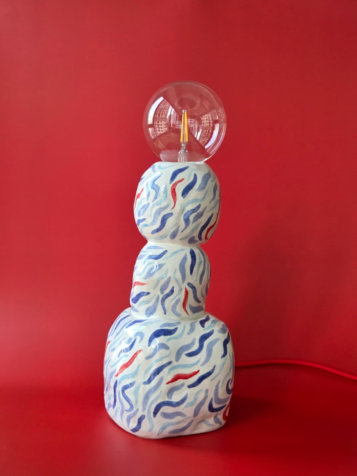 Drift, a stacked stoneware lamp hand-painted with blue and red marks and topped with a clear glass globe against a red backdrop