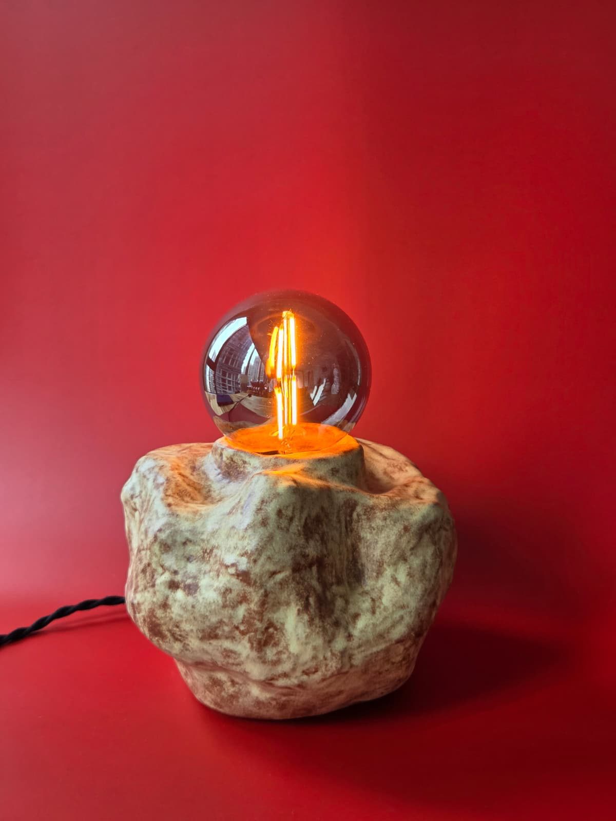 Illuminated Erratic stoneware lamp with a boulder-like form and glass globe against a red backdrop
